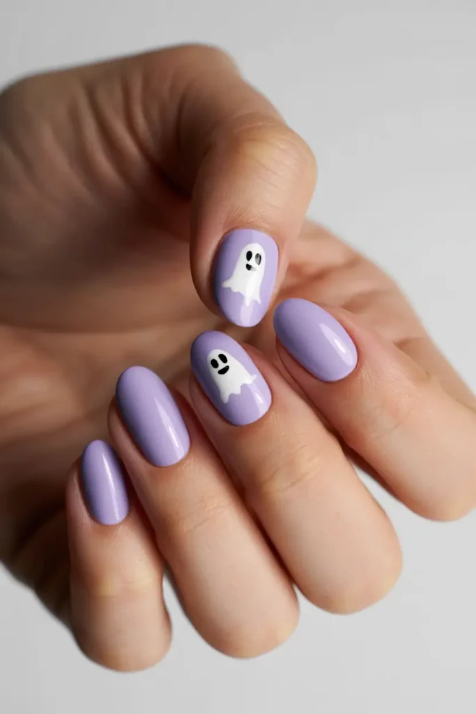 Close-up of hands with short almond nails painted soft lavender. Tiny white cartoon-style ghost nail art with black eyes. Bright, clean lighting, minimal background.