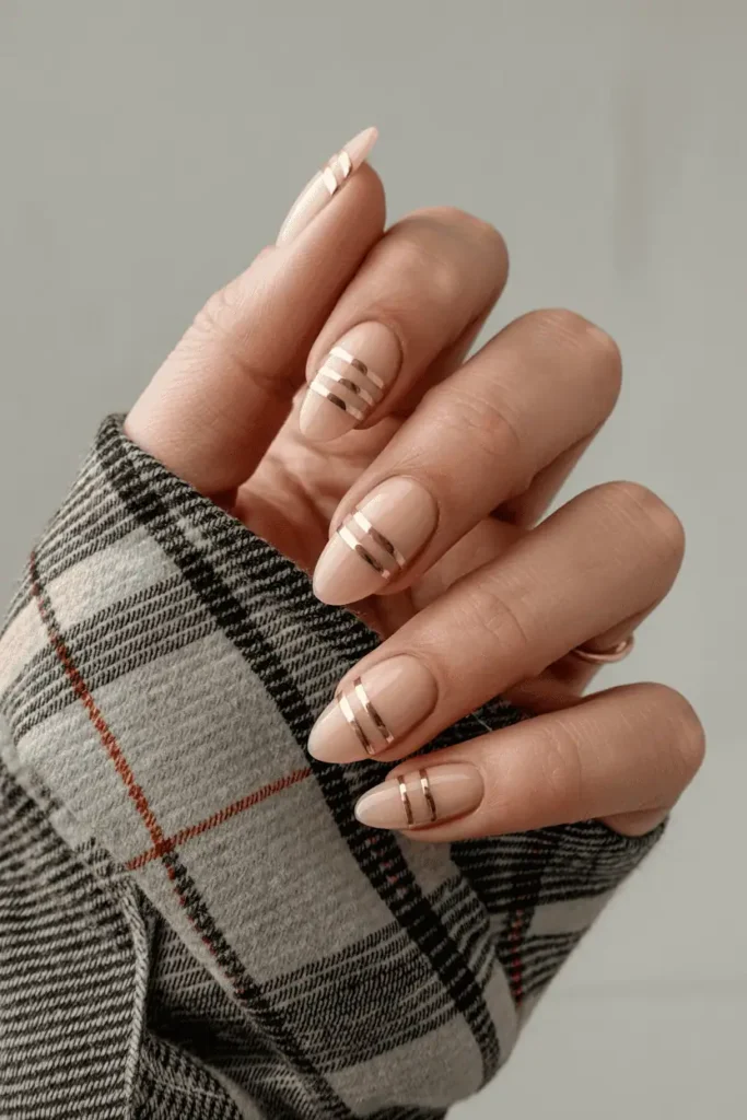 Nude almond nails with thin metallic gold striping, styled with plaid fabric background.