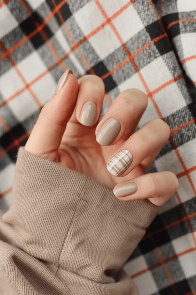Short nails with beige and white plaid pattern accents, paired with solid taupe nails. Fall aesthetic background with plaid fabric.