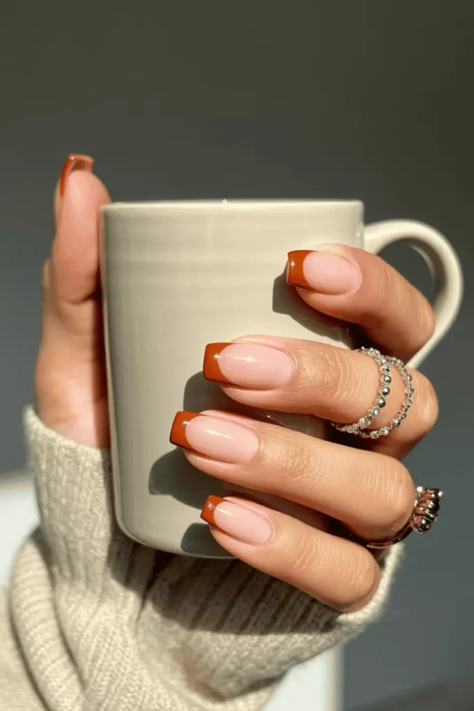 Nude nails with burnt orange French tips, styled holding a cozy coffee mug.