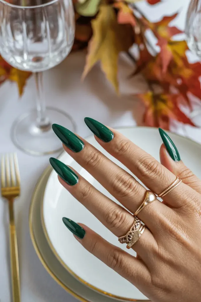 Glossy emerald green stiletto nails styled with gold rings, elegant autumn table setting.