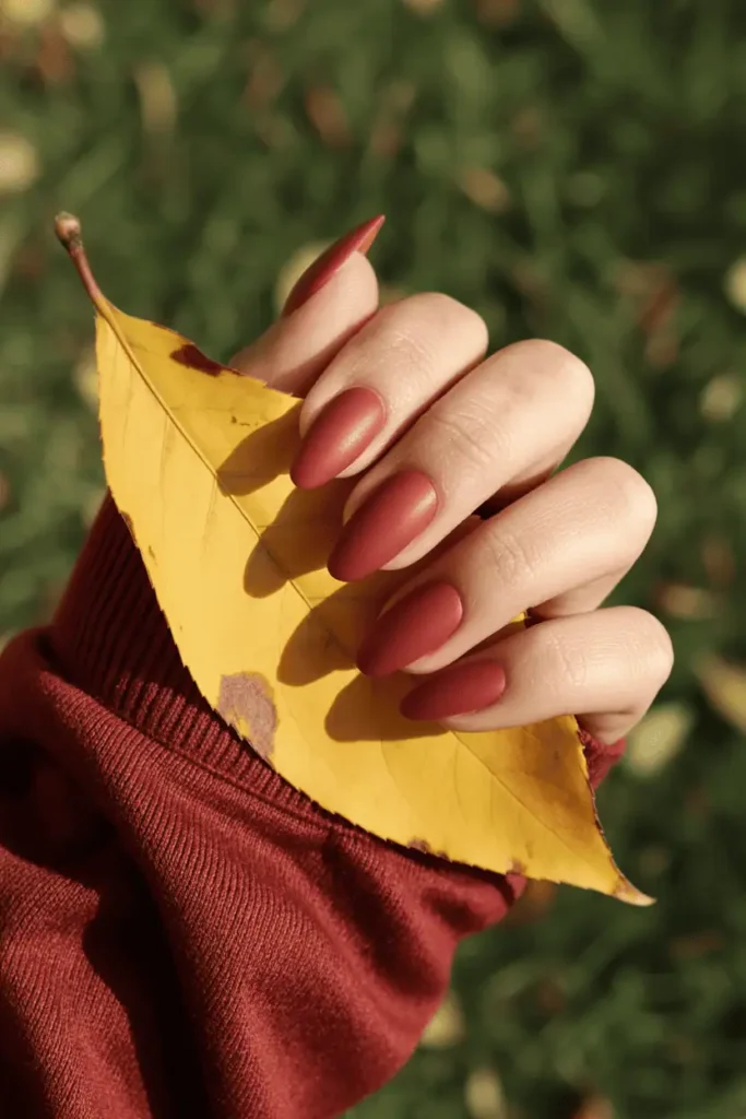 Matte rusty red almond nails holding autumn leaves, styled outdoors in natural light.