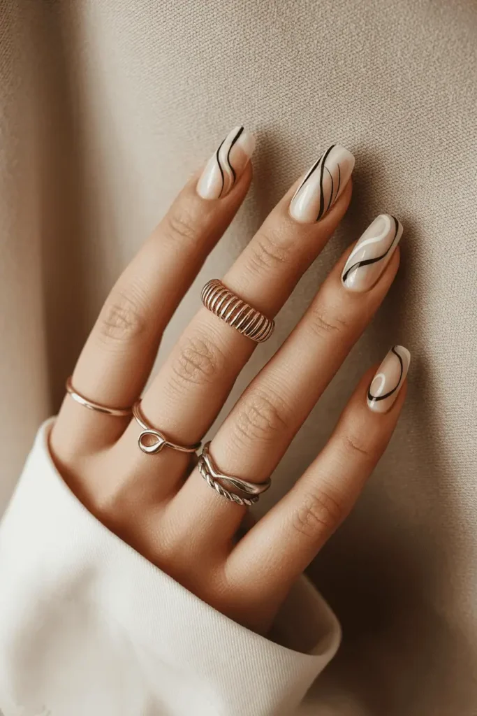 Short nude nails featuring thin black and white abstract lines. Styled with minimalist rings and neutral fabric backdrop.