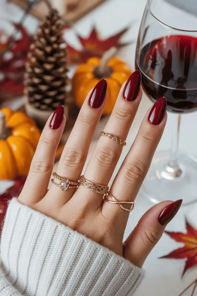Glossy burgundy almond nails styled with gold rings, cozy autumn background with wine glass.