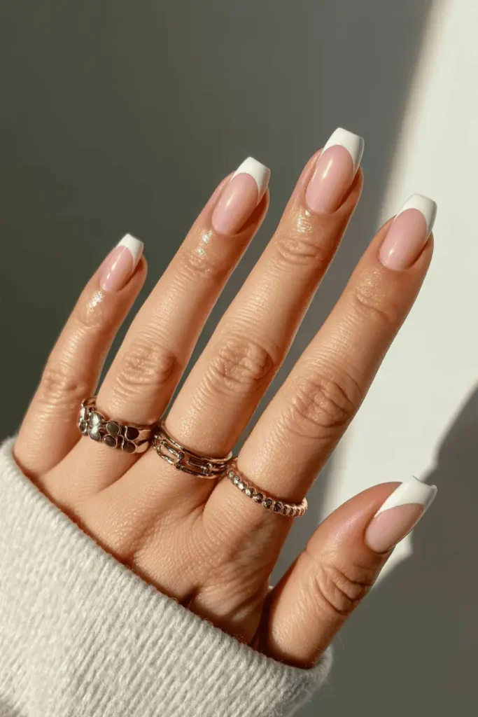 Short nails with soft white French tips on sheer pink base, glossy finish. Styled with gold rings, neutral background, natural light.