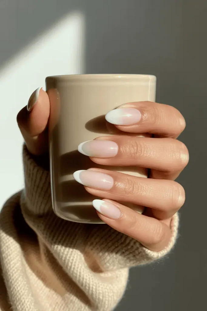 Soft almond-shaped nails painted in milky white with a creamy translucent finish. Styled holding a coffee mug on a beige sweater background, warm daylight.