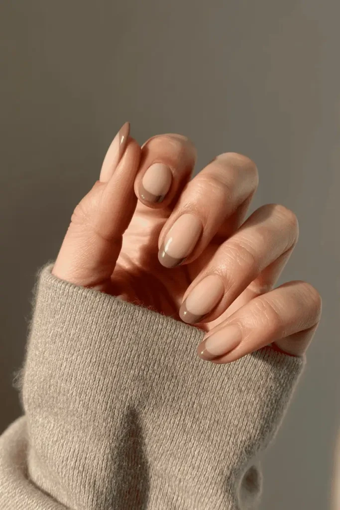 Nude base with soft taupe French tips on short almond nails. Minimal background with beige tones and soft focus.