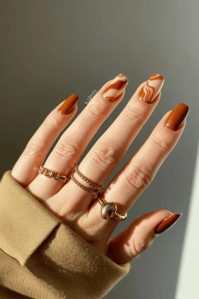 Close-up of caramel and cream marbled nails with a glossy topcoat. Styled on hands wearing gold rings and a tan coat sleeve.